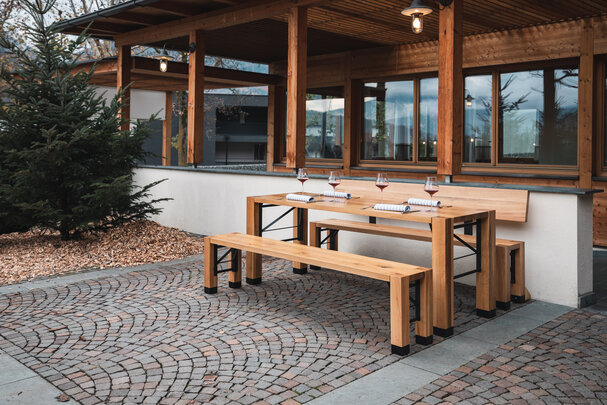 Die Designgarnitur Lago steht vor einem Gebäude. Eine Bank mit Lehne und eine ohne Lehne, der Tisch ist gedeckt.