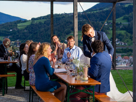 Das Foto zeigt eine Hochzeitsfeier. Braut, Bräutigam und Gäste sitzen alle auf Bierzeltgarnituren.
