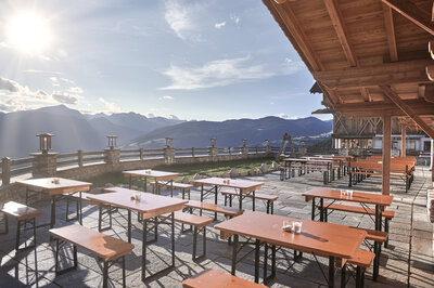Auf einer Terrasse einer Hütte stehen kleine Bierzeltgarnituren in  der Sonne.