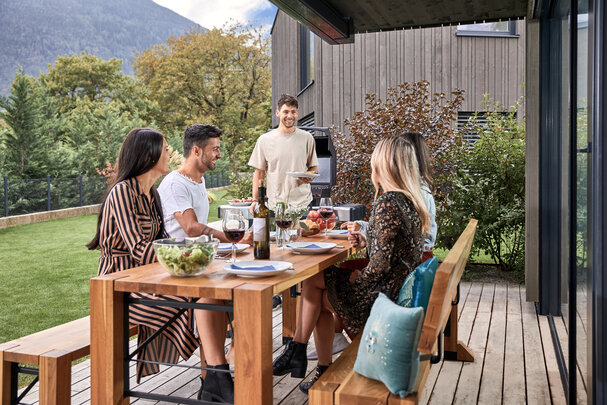 Es findet ein Grillnachmittag im Garten statt und 4 Personen sitzen bereits auf der Designgarnitur Lago und ein Mann kommt mit einem Teller auf sie zu.