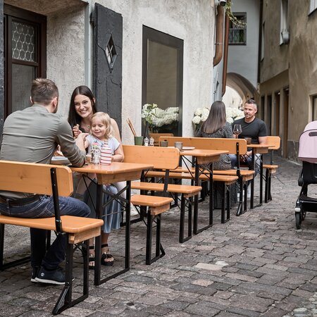Several people sitting on small beer garden table sets in an alley near a restaurant.